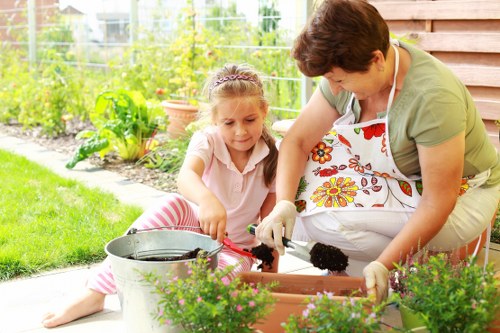A woman and a young girl are working together in a well-maintained garden outside a house, engaging in gardening activities such as planting or potting in a rectangular garden bed. The woman, wearing a floral apron and gardening gloves, is leaning over the garden bed, handling soil or plants, while the girl, dressed in a light pink top and striped leggings, sits on the grass nearby holding a garden tool. In the foreground, there are vibrant flowering plants in terracotta pots and a metal bucket, adding natural colour and texture to the scene. The garden features a lush green lawn, bordered by a variety of plants, bushes, and young trees that provide a natural backdrop, with some flower beds visible along the garden edge. The weather appears bright and mild, supporting outdoor gardening activities. This outdoor space exemplifies a typical cultivated garden suitable for regular maintenance and landscaping, reflecting professional gardening services such as those offered by Lawn Mowing Manor Park, who support garden care and outdoor improvement in the local area.